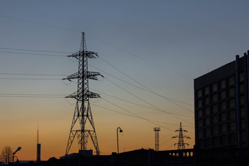 Fototapeta premium Silhouette of a pylon of a high voltage power line and a plant in the evening light of the setting sun. Electricity distribution, industry and manufacturing. Industrial landscape. Voltage transmission