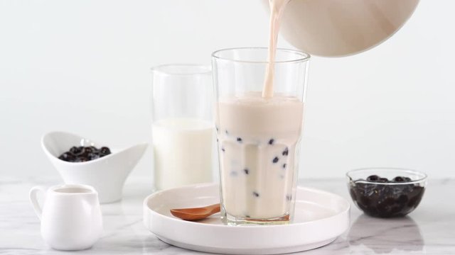 Pouring milk tea in glass cup with tasty popular taiwan tapioca pearl bubble on bright marble table and white tray, homemade concept. 4K video shot.