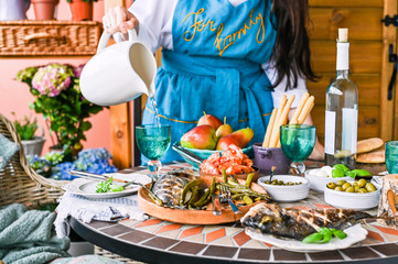 Fried fish in a dish in female hands. Dining table with different food and snacks. Summer lunch in the open air. Photo in bright colors. Woman in the frame at the dinner table.