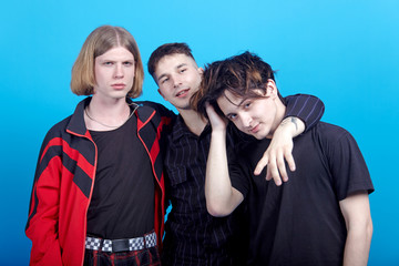Fototapeta premium Three young handsome men stand embracing. Gay relationship or close friendship. Indoors, studio portrait, copy space, blue background.
