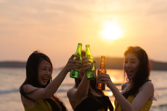 Candid Of Young Attractive Three Asian Girl Group Of Friends Cheers Holding Green Bottle Beer Drinking For Celebration Party At Sea Beach In Evening Sunset Or Silhouette Concept. Asian Tourism Moment.