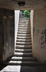 Ascending passage from underground area with contrast sunlight upstairs, weathered steps and narrow dirty concrete walls