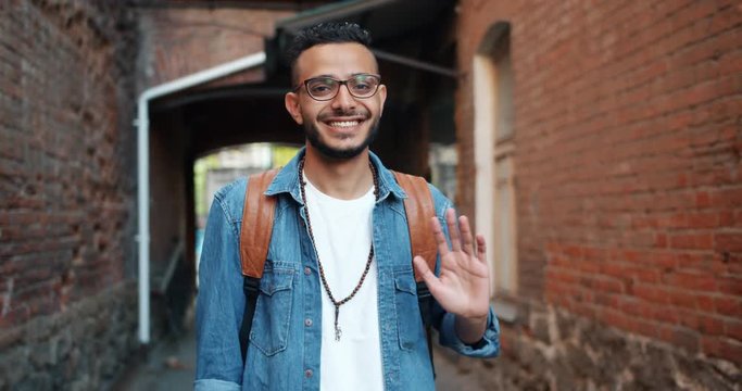 Portrait of friendly cheerful Arabian man in stylish clothing waving hand outdoors smiling looking at camera. Hand gestures, people and greeting concept.