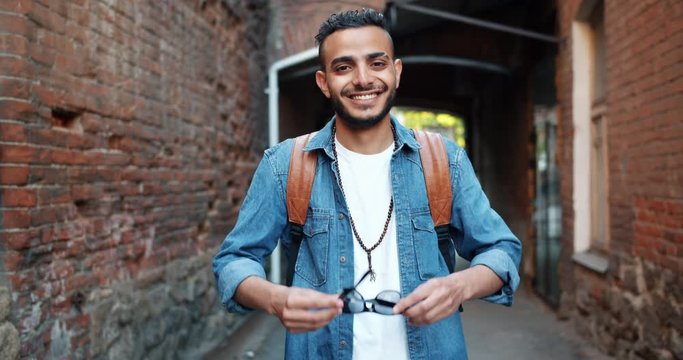 Portrait of good-looking middle Eastern man taking off sunglasses smiling standing outdoors in the street in city with hapy face. Youth and style concept.