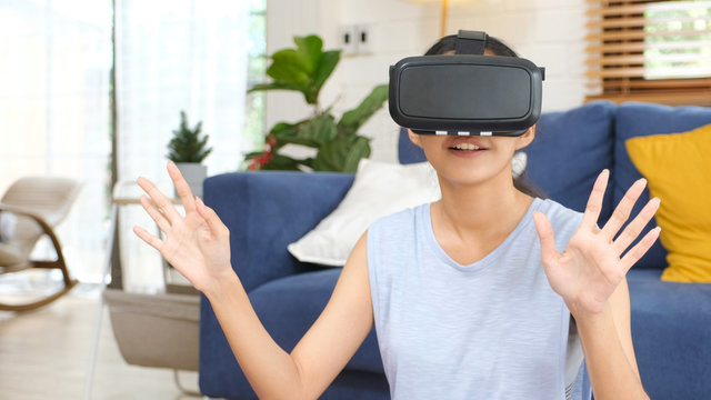 Young beautiful asian woman exciting in VR headset looking up and trying to touch objects in virtual reality at home living room, people leisure technology lifestyle