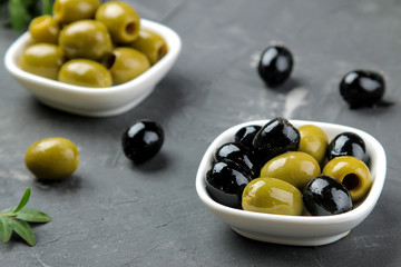 Green and black olives in a white ceramic bowl with leaves on a dark graphite background. close-up