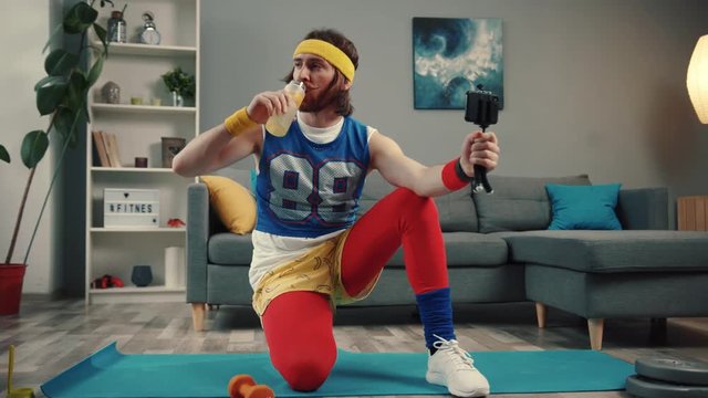 Confident male blogger in retro outfit drinking water after exercise recording a video for sports vlog at home.