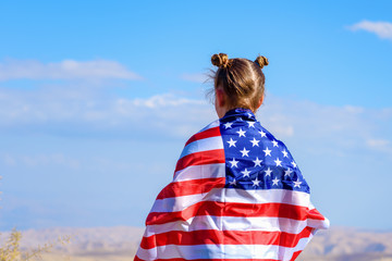 American flag. Back view little patriotic happy girl wrapping in usa national flag on blue sky background. National 4 july. Memorial day.