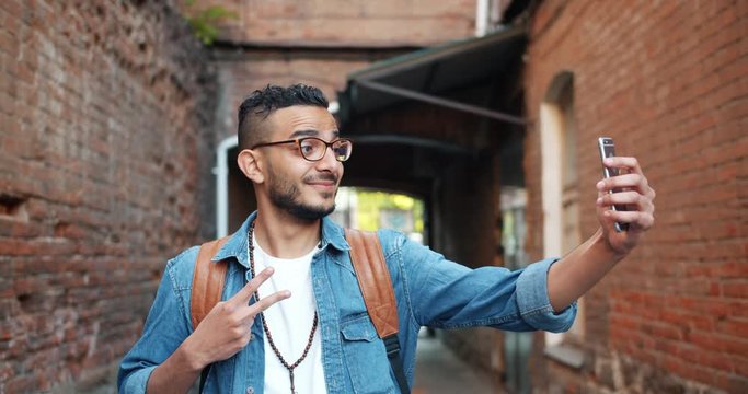 Joyful African American man is taking selfie with smartphone camera outdoors showing thumbs-up and v-sign hand gestures posing and smiling in the street.