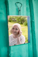 Attractive senior blonde woman looking at herself in mirror sitting outdoor at summer garden