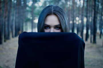 Vampire Woman in a gloomy forest. Beauty Sexy Vampire Girl covered her face with a cloak . Sexy Witch. Blue eyes and Wicked look. Attractive model girl in Halloween costume and make up. - obrazy, fototapety, plakaty