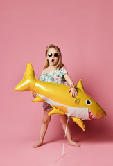 little girl, dressed in a summer dress, wears a balloon in the shape of a yellow shark-fish,...