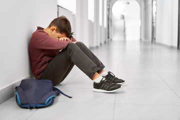 Sad school boy sitting alone on floor at corridor.