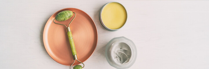 Facial beauty product jade face roller top view of massage tool with jars of shea butter and natural clay banner panorama. Skincare wellness cosmetic therapy Luxury treatment.