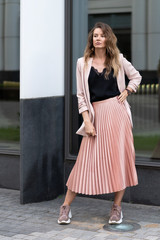 model posing in coral color clothes. Pleated long skirt, jacket, sneakers, black blouse. wavy hair below shoulders. Young beautiful european woman stands on the street near the glass wall.