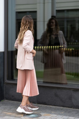 girl looks in a glass window. Peach jacket, coral skirt, pleated, sneakers. Fashion clothes. Summer day in the city.