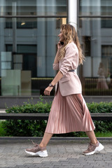 the girl is walking down the street and talking on the phone. Women's jacket and pink skirt pleated skirt. Modern look. Bench and high black glass showcases on background. Summer day.