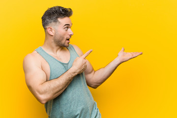 Young fitness man against a yellow background excited holding a copy space on palm.