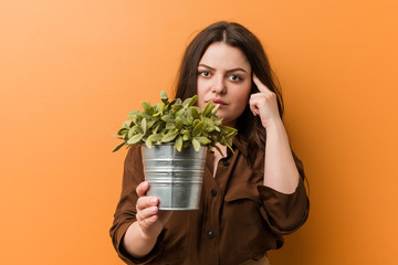 Young curvy plus size woman holding a plant pointing his temple with finger, thinking, focused on a task.