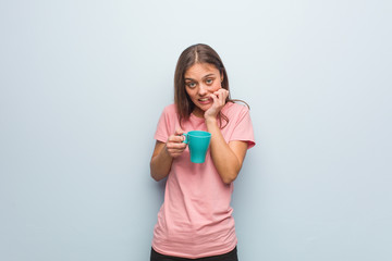Young pretty caucasian woman biting nails, nervous and very anxious. She is holding a mug.