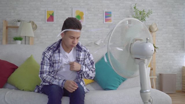 Young asian man sitting in front of a working fan, the concept of heat