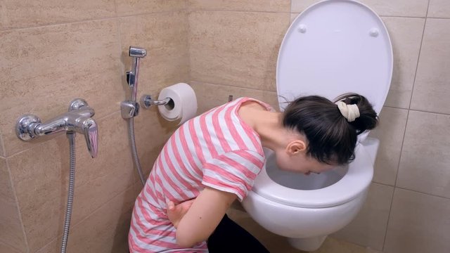 Sick young woman is vomiting in toilet sitting on the floor at home, food poisoning symptom, side view.