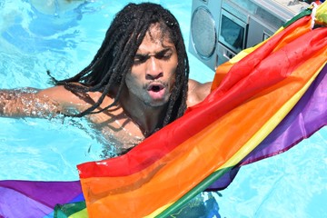 A young african-american man with dreadlocks is refreshing in a pool listening music on a vintage cassette from which hangs a rainbow flag. International LGBTI pride day celebration concept
