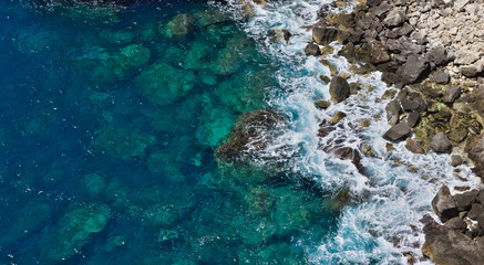 Aerial top view of crystal clear blue waves hitting rocky beach creating white foam.