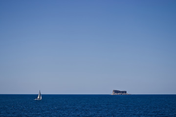 Distant Ship sailing across dark blue ocean towards small rocky island against a blue sky.