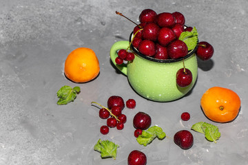 Sweet cherries in a green mug spattered with drops of cool water, next to a gray background a scattering of berries and apricots. 