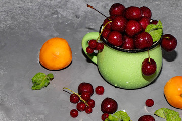 Sweet cherries in a green mug spattered with drops of cool water, next to a gray background a scattering of berries and apricots. 