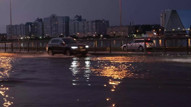 Car traffic with big splashes on the flooded city street. Large puddle on the asphalt road at night. Road after the rain. Emergency - water drains cannot cope with heavy rain, deluge.
