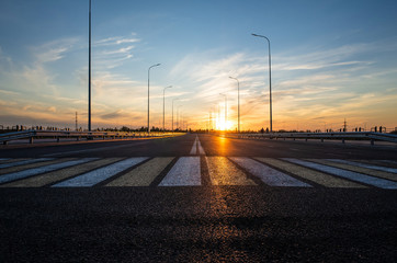 Fototapeta na wymiar Road with a pedestrian crossing on a new asphalt with a shallow depth of field, blurry background and variable focus. Sunset, warm shades. Road construction concept. Place for text.