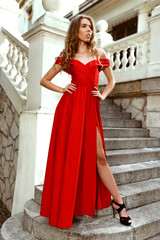 Classy woman in long red dress and black high heels, staying on stairs of palace. Concept of elegance.