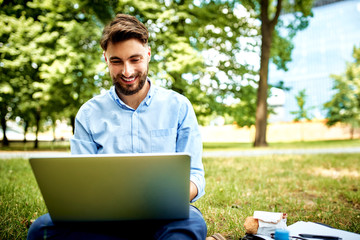 Young smiling businessman sitting on grass in park during lunch and working on laptop