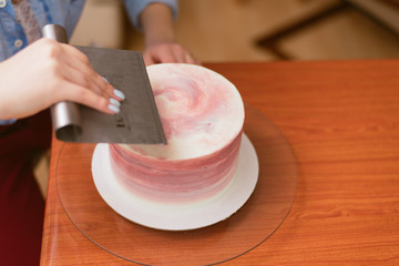 A lovely girl making a cake in a bakery. The girl smoothes the cream on the cake. White cake on a wooden table.