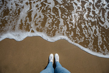 Woman feet view with jeans from above at the beach with sand and wave coming in the frame