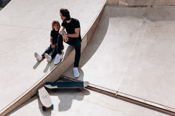 Happy father and his son dressed in the casual clothes are sitting and laughing at the slide in a skate park next to the skateboards at the sunny day