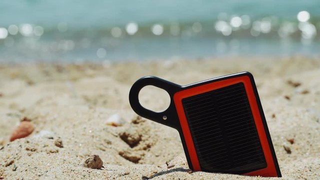 Portable solar charger power bank on the beach. Day view of small orange and black frame sun charger with black panel partly submerged on a sandy beach, with blurred sea background on a sunny day.