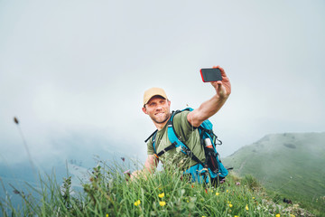 Smiling backpacker man taking selfie picture using smartphone and showing Thumbs Up during walking by the foggy cloudy weather mountain range .  Active sport backpacking healthy lifestyle concept.