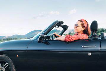 Young smiling cheerful female driving retro style convertible car at sunny day time - obrazy, fototapety, plakaty