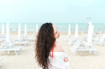 A woman in a red dress and curly hair is standing on the beach. A man with happy emotions. Walk along the sea promenade. Travel and tourism in the summer. Copy space