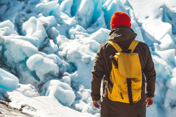 Fototapeta na wymiar Traveler man with a yellow backpack wearing a red hat standing on a rock on the background of a glacier, mountains and snow. Travel lifestyle concept. Shoot from the back