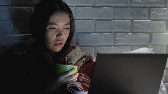 Sick woman covered in blanket drinking hot tea at night and working on laptop