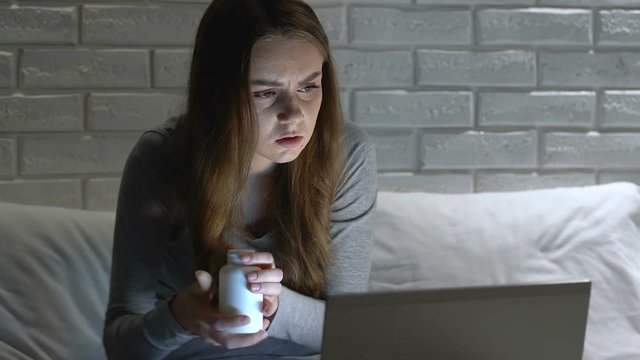 Nervous girl using laptop, taking pills at night, searching for diagnosis online