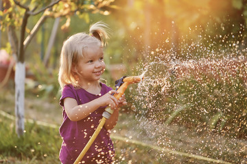 Cute little girl watering flowers in the garden at summer day. Child using garden hose on sunny...