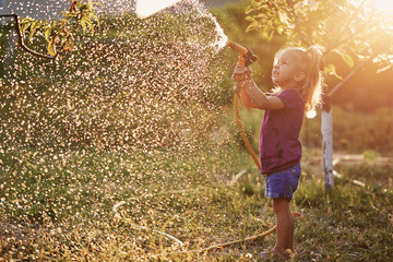 Cute little girl watering flowers in the garden at summer day. Child using garden hose on sunny...
