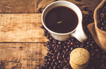 Black coffee In the cup white have roasted coffee beans In a cloth bag sack and dispersedly are on table, have milk cookies, place stacked 5 pieces, all are placed on a wooden table.