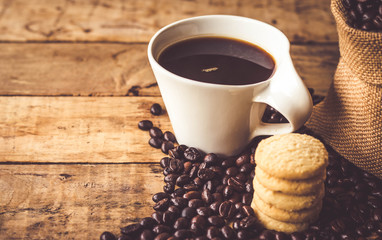 Black coffee In the cup white have roasted coffee beans In a cloth bag sack and dispersedly are on table, have milk cookies, place stacked 5 pieces, all are placed on a wooden table.