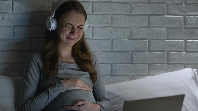 Crying pregnant woman in headphones watching melodramatic movie on laptop